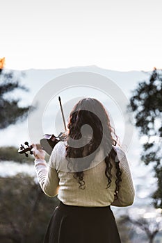 Brunette female violinist seen from back playing violin outside in the mountain. Vertical