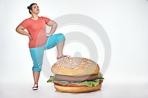 Brunette, funny, plump woman put her leg to a huge sandwich