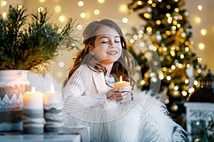 A brunette gilr in front of fur-tree and fireplace with candles and gifts. A girl making a wish. Christmas eve.