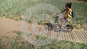 Brunette girl with a backpack rides a bicycle along a path of wooden flooring in a park on the shores of the Gulf of