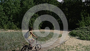 Brunette girl with a backpack rides a bicycle along a path of wooden flooring in a park on the shores of the Gulf of