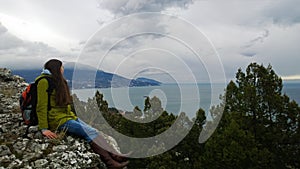 Brunette girl with a backpack wearing a coat and boots sitting on a rock and looking towards the sea and mountains.