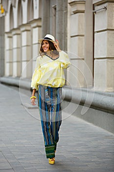 Brunette girl dressed in yellow in white peas blues, striped pants and hat walks in the city street on a sunny day