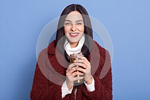 Brunette girl dresses white warm sweater and faux fur coat, posing isolated over blue background, holds coffee in thermo mug,