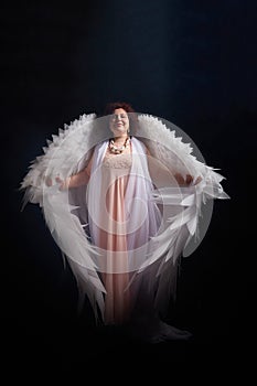 A brunette girl in an elegant dress and with white angel wings on a black background. Model, actress or dancer posing in