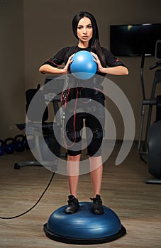 Brunette girl in EMS suit stands on bosu ball and holds blue ball. Glow effect