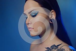 Brunette girl with a flower tattoo on her shoulder and tunnels in her ears in beautiful blue light. Woman with blue make-up,