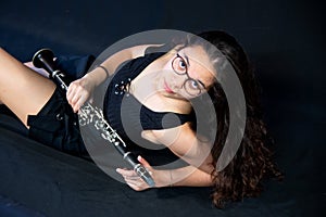 A brunette girl, with glasses, lying laughing and holding her clarinet music instrument isolated on a black background