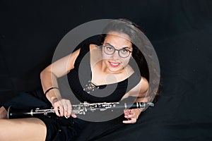 A brunette girl, with glasses, lying laughing and holding her clarinet music instrument isolated on a black background