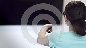 brunette girl with glasses sits on the sofa in front of the TV and switches channels, back view, cinematic shot