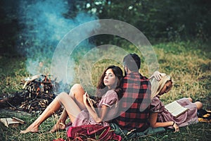 Brunette girl leaning on her boyfriend while reading book. Turn back guy looking at campfire. Young couple in love