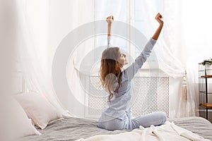 Brunette girl in the light-blue pajama stretches her arms up sitting on the canopy bed next to the window in the cozy