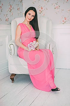 Brunette girl in pink dress sitting white armchair with bouquet of flowers in their hands