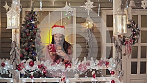 Brunette girl in santa claus hat sending greeting sms using smart phone a porch