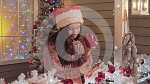 Brunette girl in santa claus hat sending greeting sms using smart phone a porch