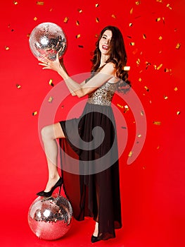 A brunette girl is standing in a black dress with gold sequins, holding a disco ball in her hands