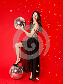 A brunette girl is standing in a black dress with gold sequins, holding a disco ball in her hands