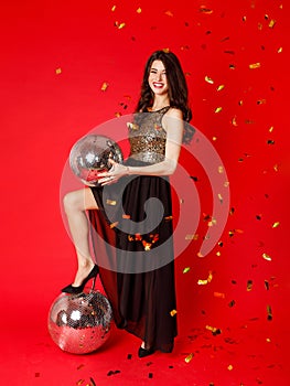 A brunette girl is standing in a black dress with gold sequins, holding a disco ball in her hands