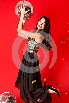 A brunette girl is standing in a black dress with gold sequins, holding a disco ball in her hands