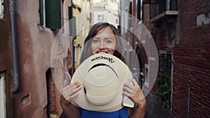Brunette girl stands on narrow Venice street, covering her face with straw hat