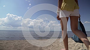 A brunette girl walks barefoot on the sand with her back to the camera towards the sea against a blue sky with white