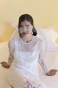 Brunette girl wearing long white lace dress sitting on bed