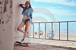 brunette girl wearing short shorts and jeans jacket standing on a skateboard and leaning on a guardrail against a