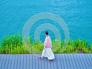 Brunette girl in a white skirt and pink jacket walking on a wooden pier path near the water