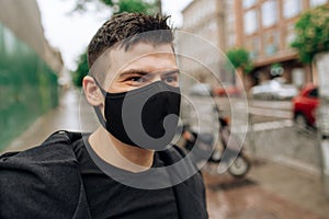 Brunette guy in a black tracksuit puts on a face mask. Black stylish face mask during a pandemic virus crown. Handmade cotton face