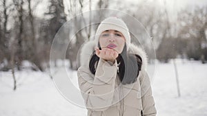 Brunette hair young women portrait with pink lips in beige down coat and hat sends an air kiss in snowy central park