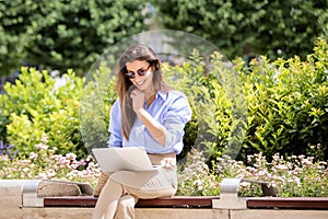 Brunette haired woman sitting on a bench in the city and using a laptop and having video call