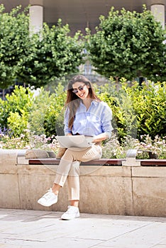 Brunette haired woman sitting on a bench in the city and using a laptop and having video call