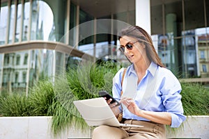 Brunette haired woman sitting on a bench in the city and using a laptop and smartphone