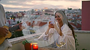 Brunette lady photographing smartphone terrace. Handsome man playing ukulele