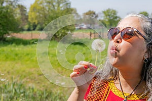 Brunette latin woman wearing sunglasses starting to blow a dandelion flower in hand