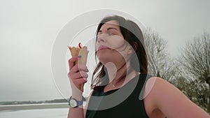 Brunette licks cone ice with pleasure. She wears black dress outside in winter. Snow on background.