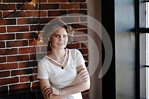 The brunette in light tight clothes posing against the background of an aged brickwork, hung with incandescent lamps.