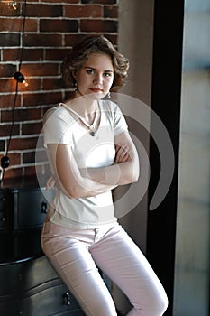 The brunette in light tight clothes posing against the background of an aged brickwork, hung with incandescent lamps.