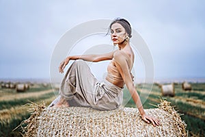 Brunette in linen pants and bare shoulders sitting on a hay bales in warm autumn day. Woman looking at camera. Behind her is a