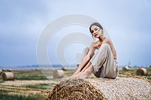 Brunette in linen pants and bare shoulders sitting on a hay bales in warm autumn day. Woman looking at camera. Behind her is a