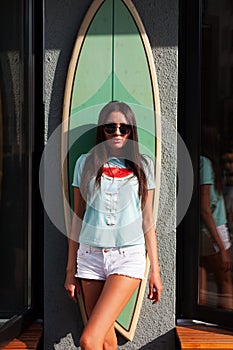 Brunette with long hair tanned leggy girl in white shorts, green T-shirt and sunglasses posing at the surf