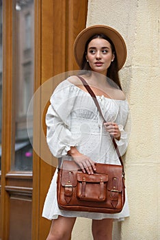 brunette with long hair wearing white romantic summer dress and beige hat posing with luxury leather bag