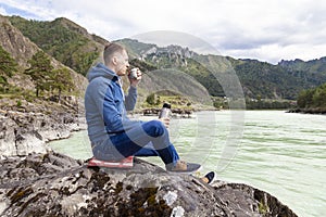 A brunette man of Caucasian appearance sits on a stone near the Katun river in the Altai mountains and drinks tea pouring into a