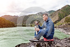 A brunette man of Caucasian appearance sits on a stone near the Katun river in the Altai mountains and drinks tea pouring into a