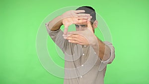 Brunette man looking through frame shape with fingers, focusing on camera. green background, chroma key
