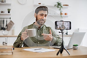 Brunette man recording vlog and holding cup in hand indoor