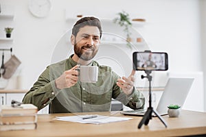 Brunette man recording vlog and holding cup in hand indoor