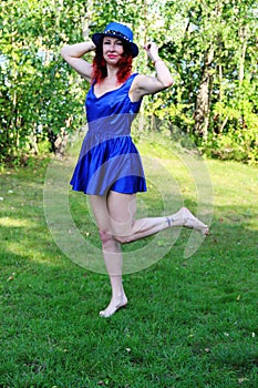 Brunette with red curly hair in a classic blue short dress and blue hat. holds his hat with his hands, jumping on one