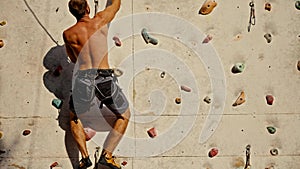 A brunette rock climber guy in gray pants with a sculpted back climbs up and climbs the climbing wall on a sunny summer