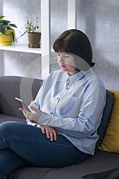 Brunette with short hair is sitting on the couch with a phone in her hands. Woman spends time aimlessly